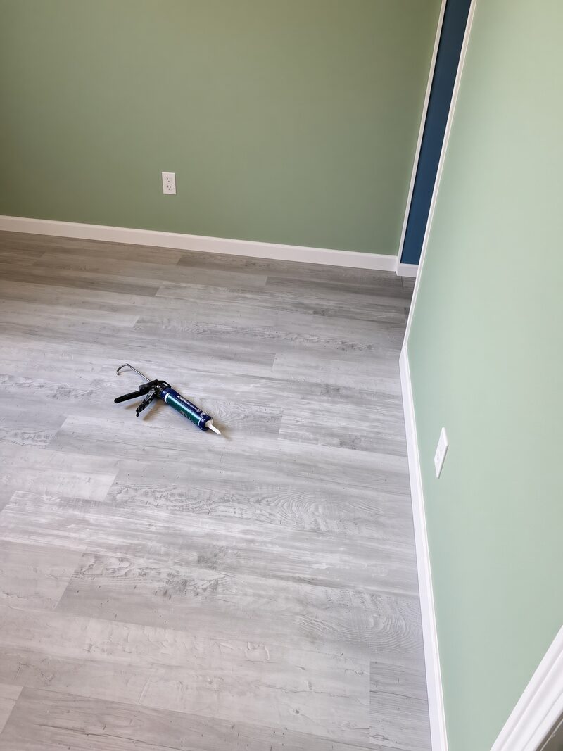 Sage green wall above grey plank flooring, caulk gun still on the floor.